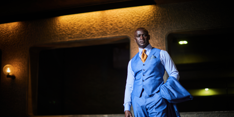 A person a blue three-piece suit with an orange tie stands indoors, holding his jacket in one hand. The background features warm lighting and a textured wall.