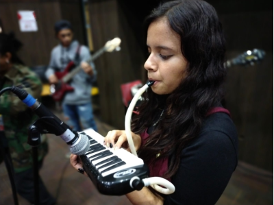 Colombian young woman playing melodica with person playing bass guitar in the background