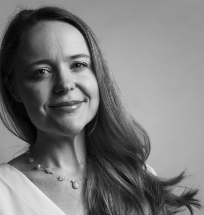 Square black and white headshot of woman looking into camera smiling, with long dark hair wearing a white top. 